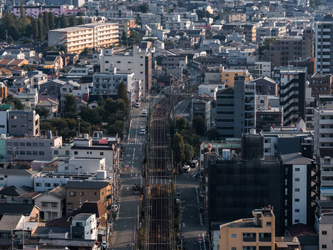 Aerial view in Osaka shows parallel railway tracks, residential blocks, solar panels, compact streets, parked cars, and warm late afternoon light with long shadows.