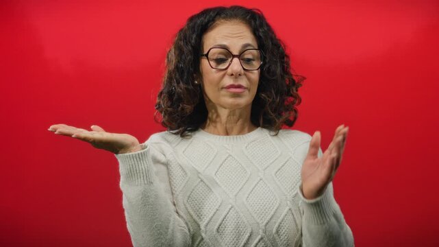 Woman with curly hair stands against a red background, wearing glasses and a sweater, extending her palms outward in a gesture, portraying choice or decision.