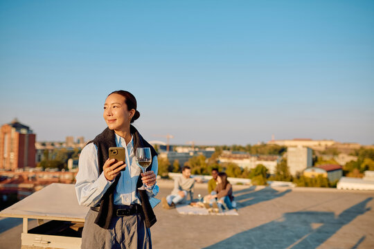 Asian woman enjoying a social gathering on a city rooftop, holding a smartphone and a glass of white wine