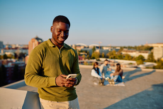 Smiling African American man standing on a rooftop, using smartphone, friends relaxing in background, enjoying urban sunset
