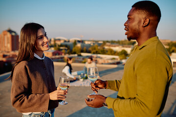 Young diverse couple enjoying sunset rooftop party, drinking white wine, talking and smiling with friends in background
