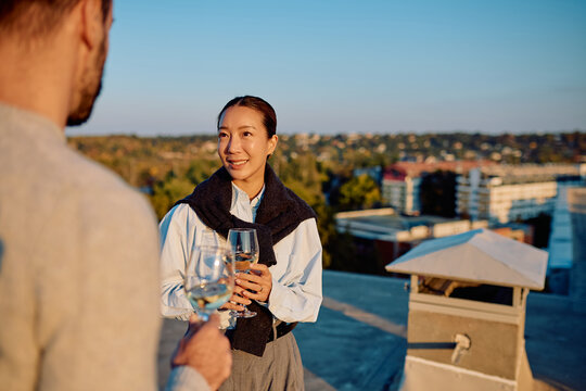 Asian woman and man enjoying romantic date on urban rooftop. Holding wine glasses and talking at sunset with city view