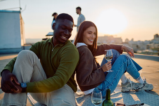 Diverse couple smiling, sitting back to back on a blanket, enjoying white wine on a city rooftop with friends at sunset