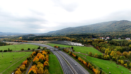 Aerial highway view through green fields and gentle hills. Autumn trees and solar panels highlight sustainable travel and countryside energy harmony. Calm landscape under cloudy sky.
