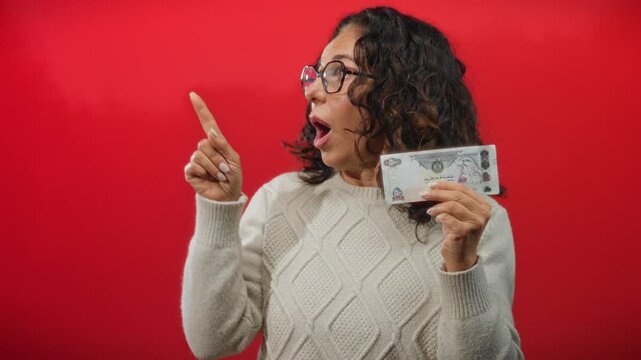 Middle-aged woman holding uae dirham banknote, surprised expression, red background, wearing glasses and sweater, signifying money decisions.