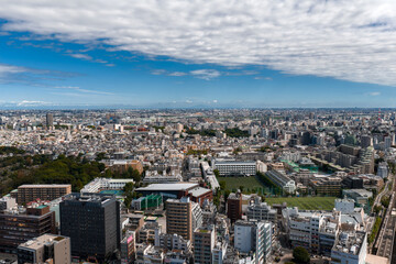 Aerial view over central Tokyo, Japan, shows dense housing, mid rise blocks, school sports fields, a domed stadium, rail and elevated roads in bright midday light under clouds.