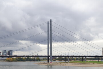 Düsseldorf Suspension/Cable-Stayed Bridge Over River With City Skyline And Cloudy Sky