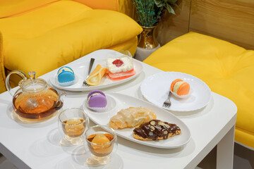 Cozy tea time setup with colorful macarons, croissants, and a slice of cake served with hot tea on white table and yellow cushions, creating a warm and inviting dessert cafe atmosphere