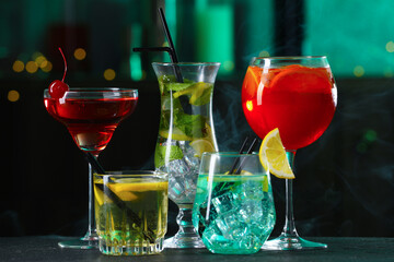 Glasses of tasty cocktails on dark table against blurred background with smoke, bokeh effect