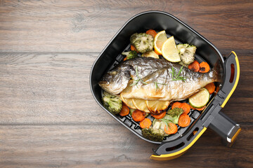 Cooked fish, lemon and vegetables in air fryer on wooden table, top view. Space for text