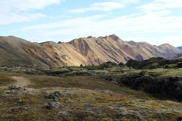 A captivating landscape showcasing a range of multicolored volcanic mountains under a partly cloudy sky. The earthy tones of brown, orange, and beige in the mountains reflect rich geological diversity