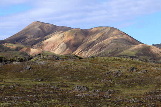 A striking landscape featuring a range of multicolored mountains in earthy tones of brown, tan, and green, set beneath a partly cloudy sky with soft blue hues