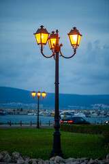 A tall, multi-headed street lamp featuring three traditional, decorative lanterns is brightly lit with warm, yellow-orange light against a twilight or blue hour sky. The black metal lamp post is situa