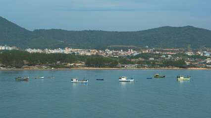 Obraz premium Coastal town scene with colorful fishing boats dotting calm blue waters. A hillside backdrop provides a scenic view of this peaceful seaside community