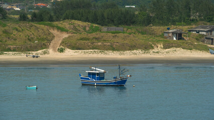 Vibrant blue fishing boat rests on calm waters near a sandy beach and grassy dunes. Peaceful coastal scene with a serene atmosphere