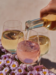 A hand pours rosé wine into elegant glasses surrounded by purple daisies in natural light. Floral summer drink scene blending freshness and celebration.