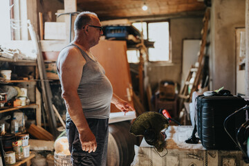 A middle-aged man in a gray tank top and glasses stands in a busy workshop filled with lumber,...
