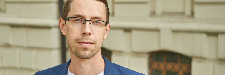 Fototapeta premium Young caucasian male with glasses and blue jacket standing outdoors in urban setting.
