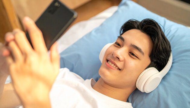 Content young Asian man enjoys digital entertainment on his smartphone while relaxing comfortably on a bed with white wireless headphones
