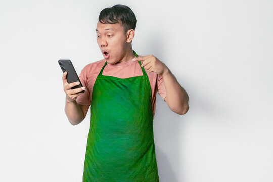 Surprised Asian man pointing and looking at his smartphone. The man is wearing a pink shirt and a green apron.