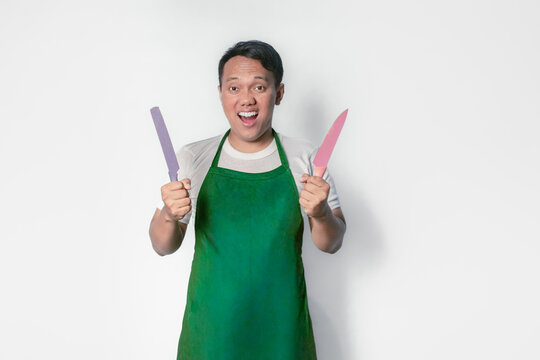 An Asian man wearing a green apron holds two knives, smiles and prepares to cook. - Powered by Adobe