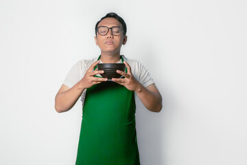 Asian man wearing green apron and glasses holding black bowl and behaving as if he is smelling food