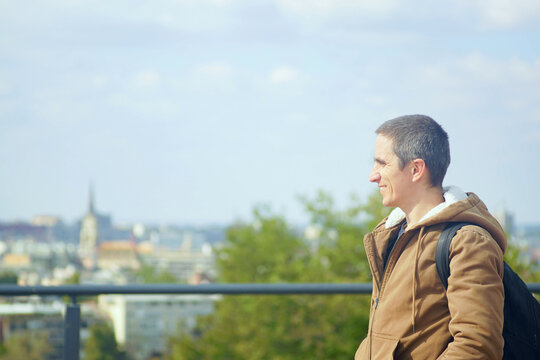 A portrait of a smiling man standing against a blurred panorama of Novi Sad. Symbolizes sincere emotions of travel, joy of discovering new places, authentic lifestyle expression, and slow tourism.