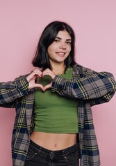 Young woman with dark hair creates heart shape with hands against a pink background in a casual...