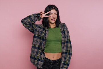 Young woman posing playfully against a pink backdrop in casual outfit while making peace sign and...