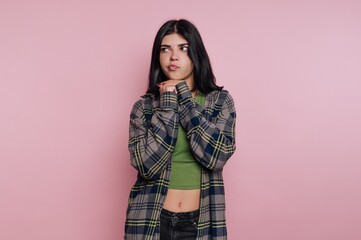 Young woman poses playfully against a pink background wearing a plaid shirt and green top