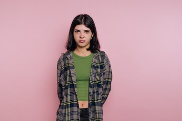 Young woman stands with serious expression in front of pink background wearing casual outfit during...