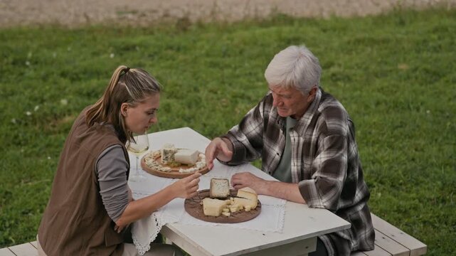 High angle shot of young Caucasian woman tasting artisan cheese made by her senior father in their family creamery, sitting at table outdoors and enjoying its exceptional flavor