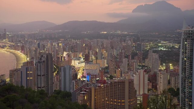 Panoramic aerial view of benidorm skyline in spain, with illuminated skyscrapers and coastline at sunset, showing urban lights turning on as dusk falls over mountains
