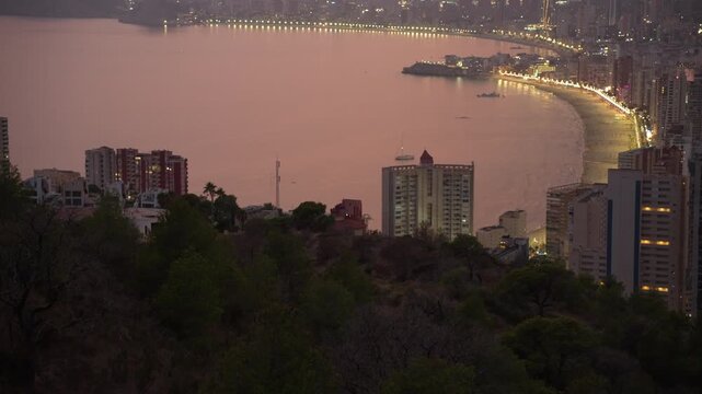 Beautiful panoramic view of the coastal city of benidorm at night, with city lights illuminating the famous beach and skyline as twilight transitions from sunset to a dark, hazy evening