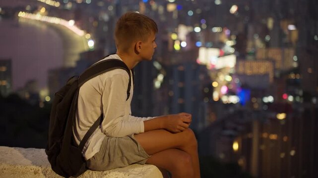 Pensive teenage boy with backpack sitting on ledge, thoughtfully contemplating vibrant, illuminated city skyline and blurred bokeh lights of urban landscape from high viewpoint