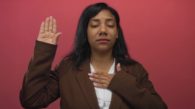 African american woman raises hand and touches chest with eyes closed in pink studio; pledge sincerity.