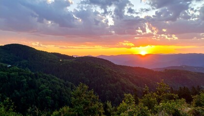 A stunning vista of rolling hills covered in lush green trees, framed by a vibrant sunset with clouds. Golden sunlight bathes the landscape