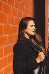 Fototapeta premium Attractive long brunette hair woman standing near brick wall. Business woman wear jacket and black bag, she look sensual, stands leaning against the brick wall, she enjoy of sunny light day.
