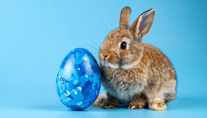 Light brown rabbit beside marbled blue Easter egg on soft blue background—evoking springtime charm and whimsical celebration.