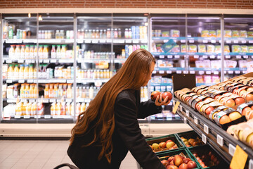 Young business long hair brunette woman girl buyer client consumer stands in shop store supermarket choosing food buying puts in basket. Woman choose fruits - apples. Healthy food.