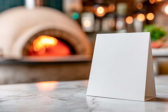 Blank tent card on a marble counter in a cozy restaurant with a wood-fired oven glowing in the blurred background.