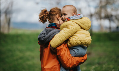 A mother is carrying her child on a spring day in a green outdoor area, showcasing family love, connection, and joy. The baby is wearing a yellow jacket, enjoying nature and bonding with the parent.