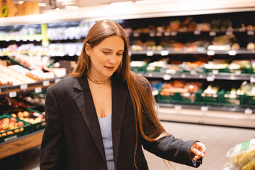 Young woman girl buyer client consumer stands in shop store supermarket choosing food buying puts in basket. Woman choose fruits pears. Business woman make shopping after work.