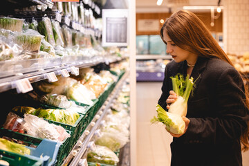 Young long hair woman in jacket chooses fennel in the vegetable section of a supermarket. Customer in a grocery store holds fennel in her hands. 