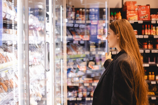 Woman in a supermarket standing in front of the freezer looking for her favorite frozen food. Woman opens the refrigerator door with a product in a supermarket. Girl chooses salmon in grocery store.
