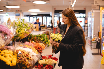 Long hair brunette woman wear jacket and choose bouquet of flowers, serious calm girl, she walk in supermarket, she is shopping after work. Garden market.