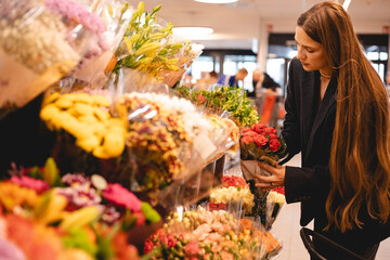 Long hair brunette woman wear jacket and choose bouquet of flowers, serious calm girl, she walk in supermarket, she is shopping after work. Garden market.