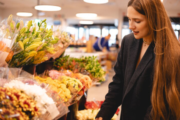 Long hair brunette woman wear jacket and choose bouquet of flowers, happy smiling girl, she walk in supermarket, she is shopping after work. Garden market.