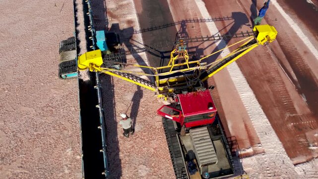 Drone top down aerial view of industrial salt harvesting equipment transferring freshly collected salt via conveyor system. Heavy machinery and workers operate at a salt production facility, showcasin