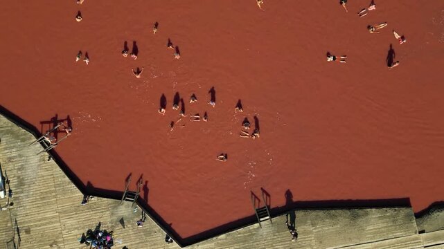 Aerial drone shot of tourists bathing and floating effortlessly in the pink saline waters of a salt lake, showing wooden platforms, mineral-rich water, and natural spa relaxation under sunlight
